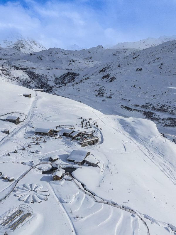 Vue aérienne de Chez Pépé Nicolas en hiver, avec ses chalets et terrasses entourés de montagnes enneigées entre Les Menuires et Val Thorens.